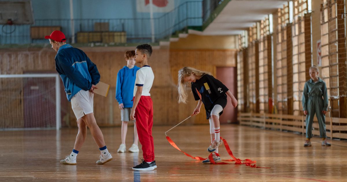 A coach with a clipboard and children with a ribbon in a wide, sunlit gym
