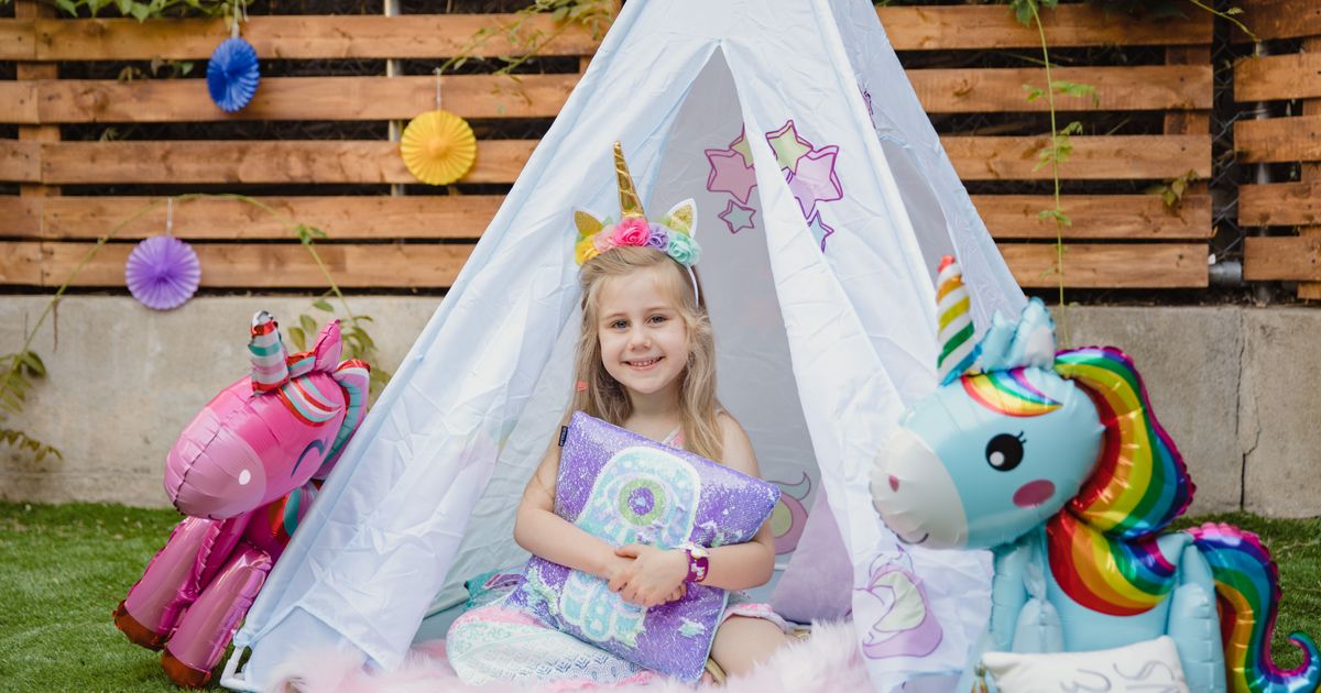 A birthday child in a unicorn headband, in a teepee between two unicorn balloons