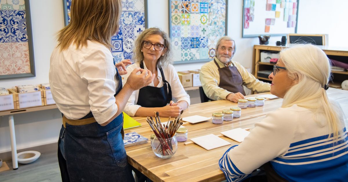 Adults round a table at a one-day tile painting workshop, listening to the instructor
