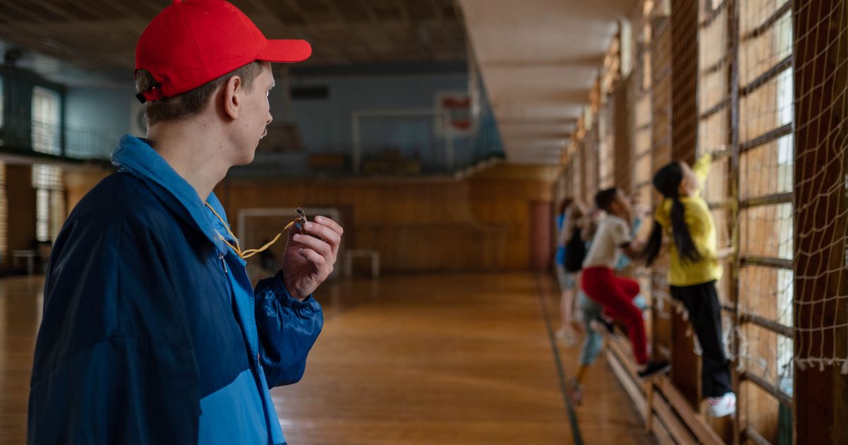 A coach with a whistle watching a children's class climb the wall bars in a gym