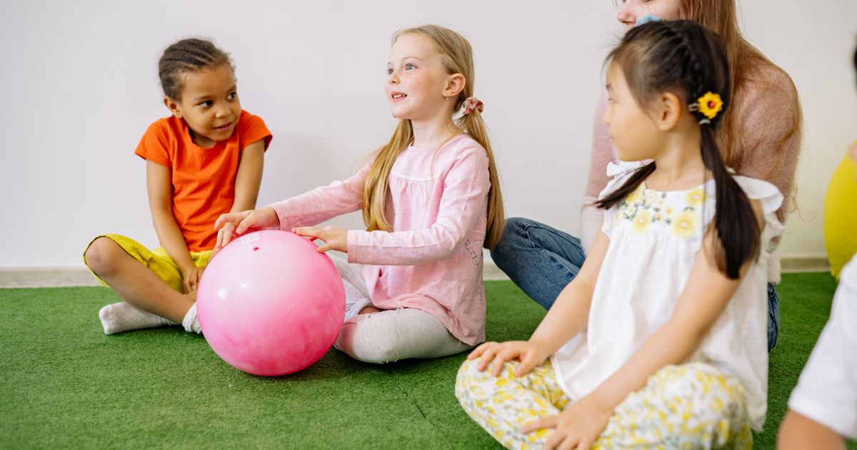 Children sitting in a circle passing a ball, with their host beside them