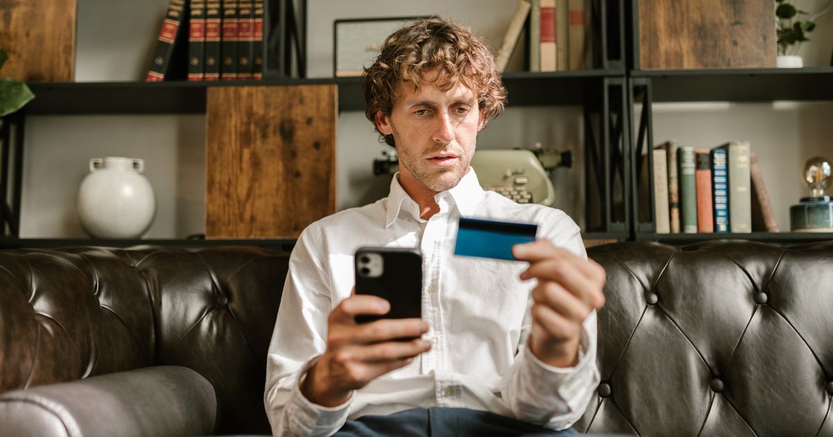 A parent on a sofa paying on their phone, a card in their other hand