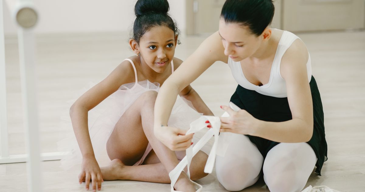 An older dancer sitting on the studio floor to help a young dancer with their shoe ribbons
