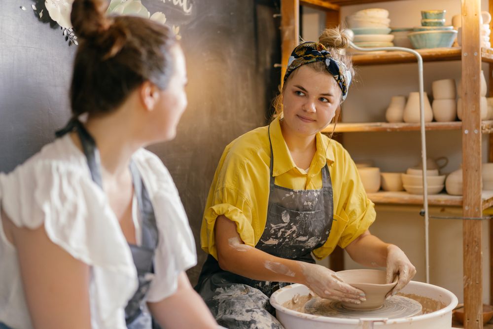 A potter shaping a bowl at the wheel while a student watches
